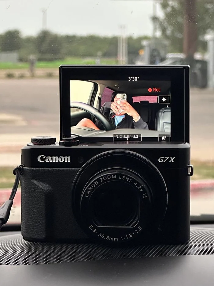 A black Canon G7 X camera on a car dashboard, its flip screen recording