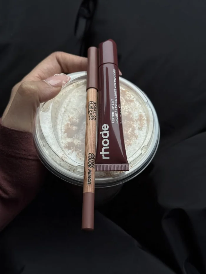 A Make Up For Ever lip pencil and a Rhode peptide lip tint held against an iced coffee